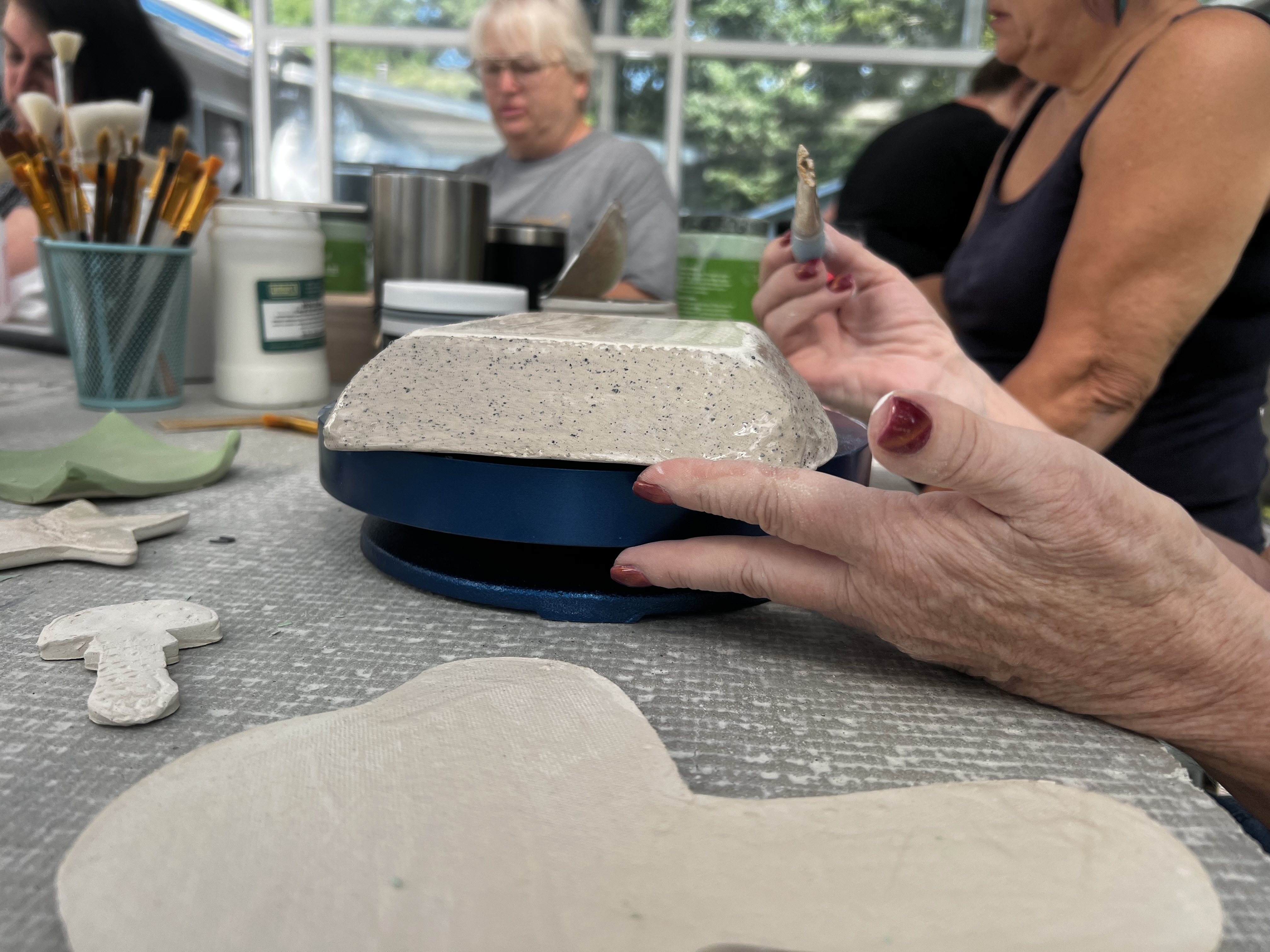 A student shapes a clay slab on a banding wheel during a hand-building class, with brushes and cut-out shapes on the table